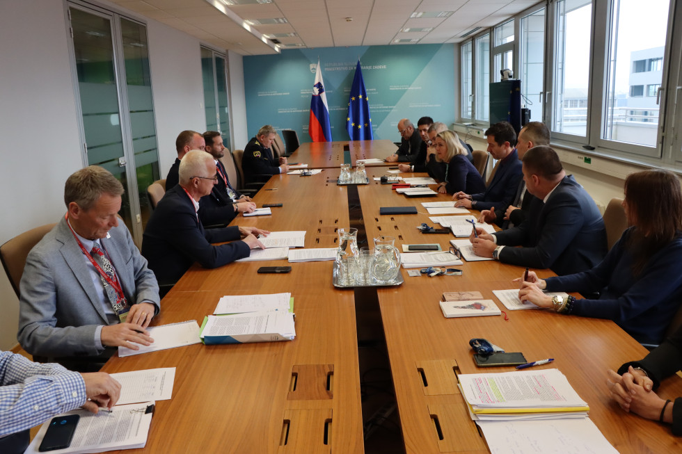 Udeleženci sestanka sedijo za dolgo rjavo mizo, pravokotne postavitve. Na koncu je modra stena, z zastavama Slovenije in Evropske unije. 