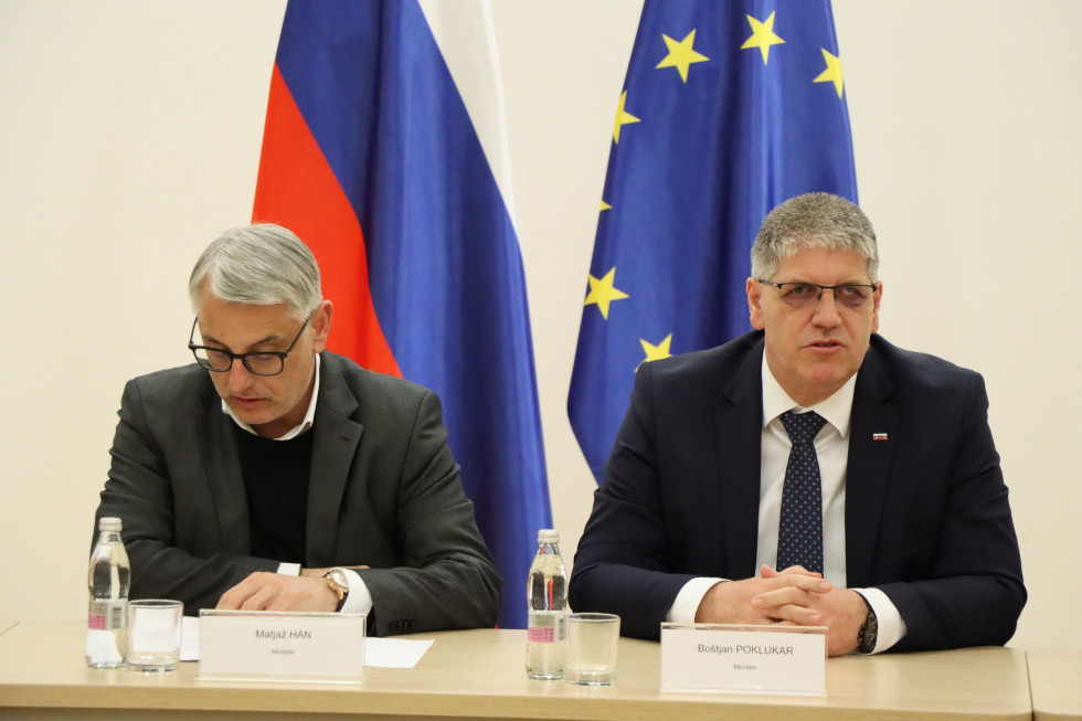 Ministra za notranje zadeve Boštjan Poklukar ter za gospodarstvo, turizem in šport Matjaž Han sedita za leseno mizo na kateri sta tablici z njunima imenoma, za njima sta vidni zastavi Slovenije in Evropske unije.