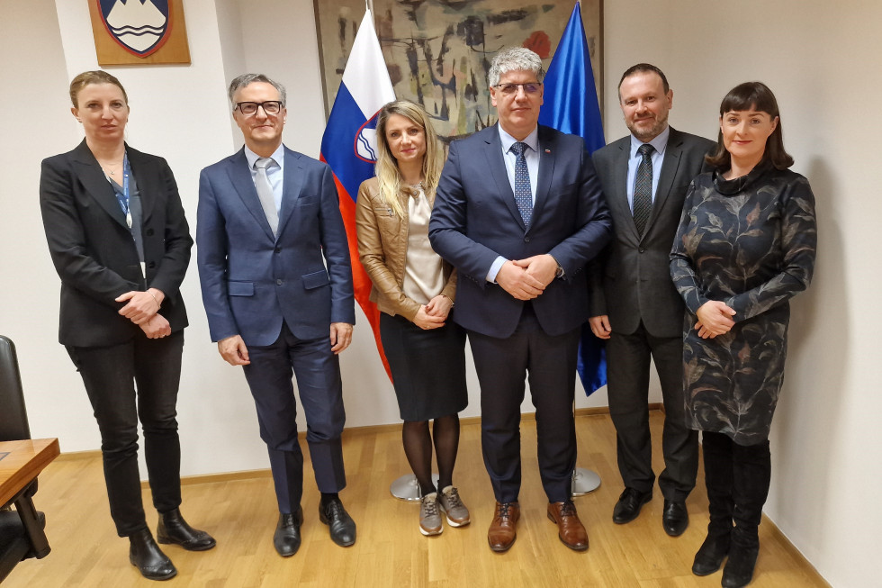 Srečanje ministra s sodelavci s poslancema italijanske in madžarske narodne skupnosti. Stojijo v sejni sobi na parketu. Za njimi umetniška slika, slovenska in evropska zastava.