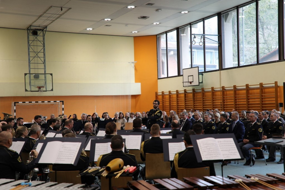 Gledalci sedijo na stolih in gledajo v člane Policijskega orkestra. Za gledalci so letveniki, nad letveniki velika steklena okna.