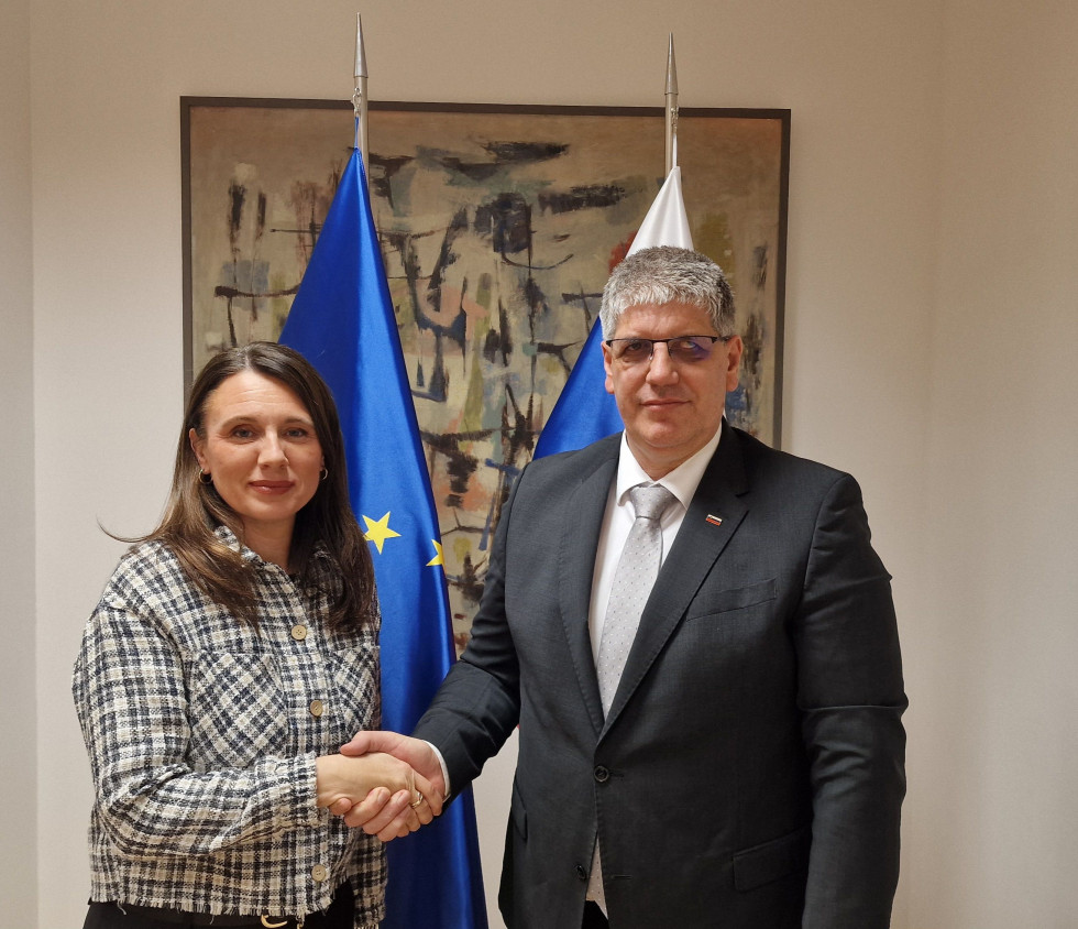 Minister Boštjan Poklukar in izvršna direktorica Agencije Evropske unije za azil Nina Gregori stojita pred slovensko zastavo in zastavo Evropska unije in se rokujeta. 