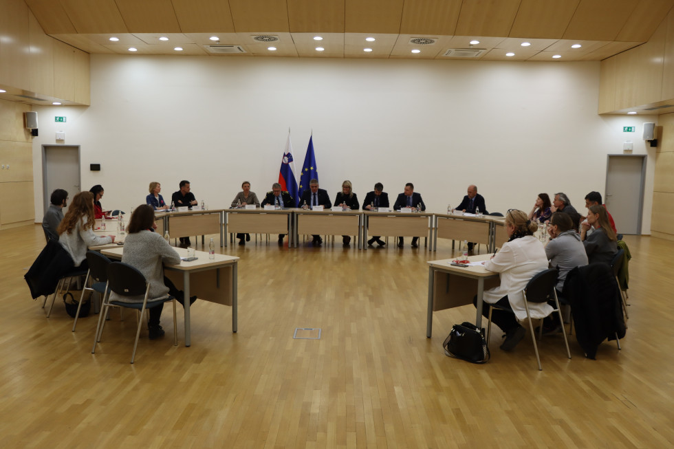 Na fotografiji so udeleženci sestanka vodstva Ministrstva za notranje zadeve in Policije s predstavniki nevladnih organizacij, sedijo za mizo v obliki črke U, drug ob drugem, nekateri pišejo, drugi poslušajo, za njimi slovenska in evropska zastava