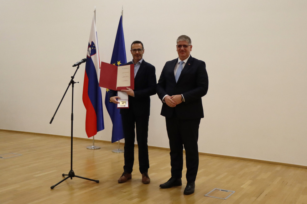 Pred zastavami stojita minister Boštjan Poklukar in dekan Fakultete za varnostne vede Univerze v Mariboru prof. dr. Igor Bernik, ki ima v roki plaketo in priznanje.