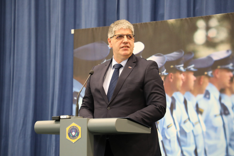Minister Boštjan Poklukar stoji za sivo govornico, za njim velik plakat, na katerem so narisani policisti, ozadje je modro.