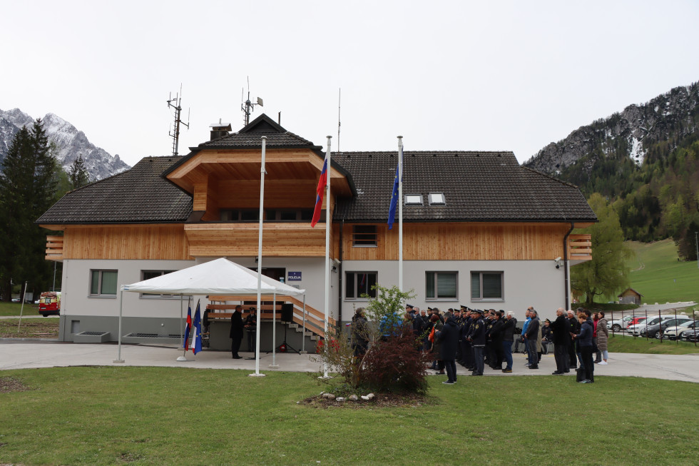 Prenovljena stavba Policijske postaje Kranjska Gora. Pogled na stavbo v alpskem stilu. Ima belo fasado, sivo streho in lesene dele med streho in fasado. Spredaj je zelenica.