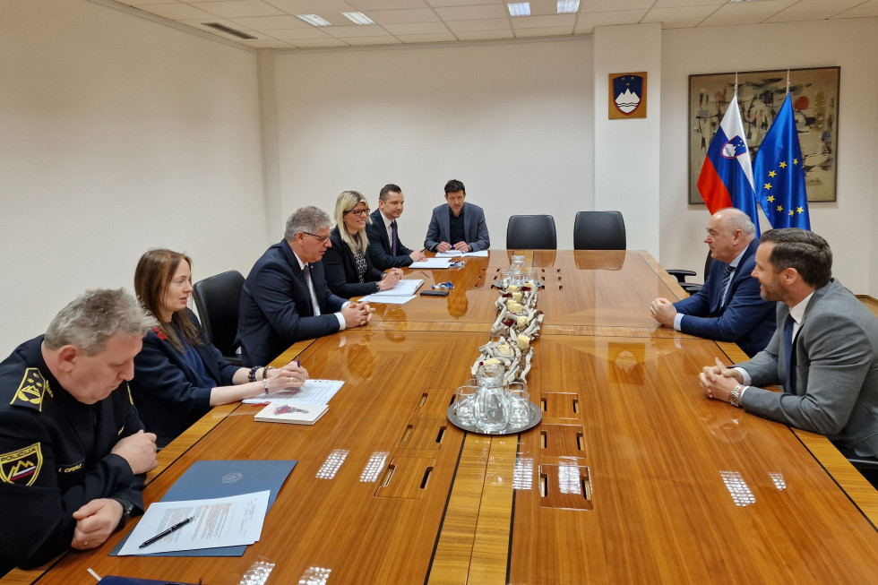 Sestanek Ministrstva za notranje zadeve z Nogometno zvezo Slovenije. Sedijo na obeh straneh podolgovate svetlo rjave lakirane sejne mize. Na mizi vrči in kozarci za vodo ter suh cvetlični aranžma. Soba ima bele stene, zadaj slovenski grb, desno zastave.