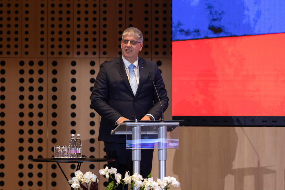 Nagovor ministra za notranje zadeve Boštjana Poklukarja. Stoji za stekleno govornico v dvorani, zadaj rjavo leseno ozadje, desno je del velikega zaslona v modri in rdeči barvi.
