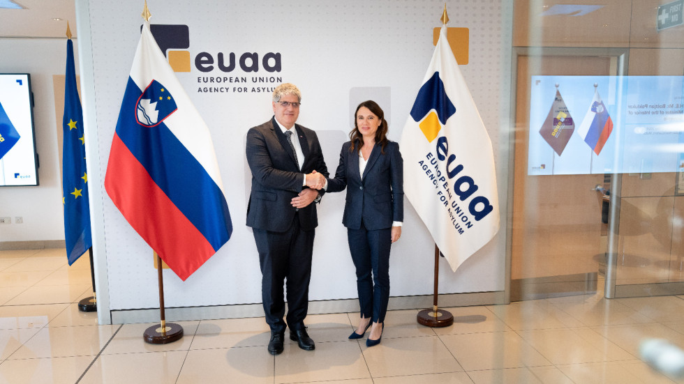 Minister of the Interior Boštjan Poklukar and Executive Director of the European Union Asylum Agency Nina Gregori stand in front of the flags of Slovenia and the Agency, the background is white and the floor is beige.