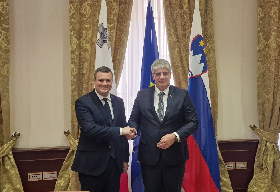 The two ministers stand in front of the flags of Malta, the European Union and Slovenia and shake hands.