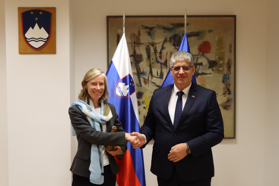 Veleposlanica Tiffany Sadler in minister Boštjan Poklukar se rokujeta, za njima so zastave in velika slika rjave barve.