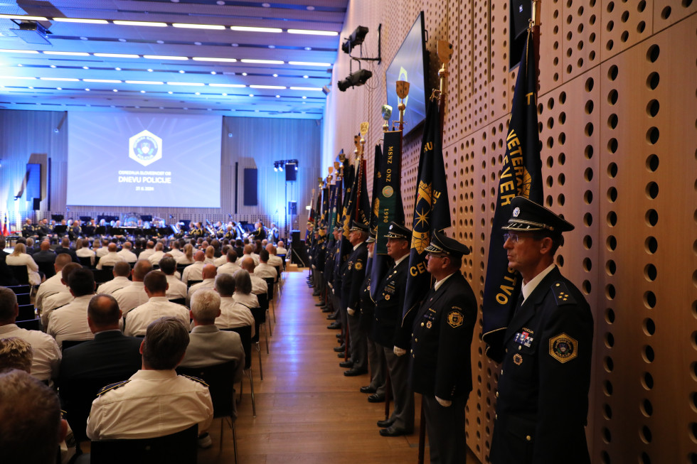 Pogled na dvorano z občinstvom in velikim odrom ter zaslonom od zadaj. Sedijo policisti v belih srajcah, ob robu dvorane stojijo policisti v uniformah.