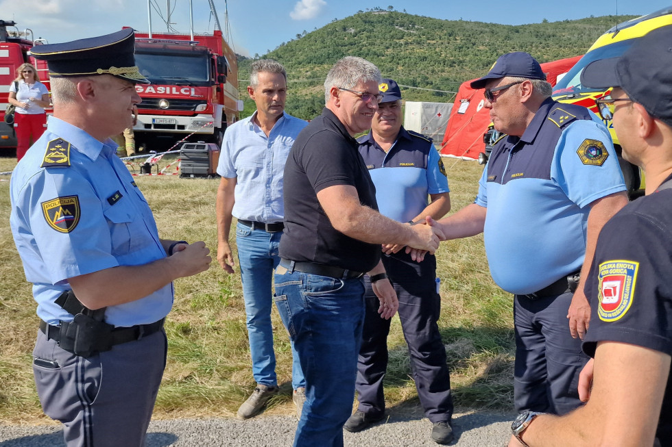 Minister Boštjan Poklukar se rokuje s policisti v uniformah. Stojijo na prostem na travi, zadaj rdeče gasilsko vozilo, v daljavi porasel hrib.