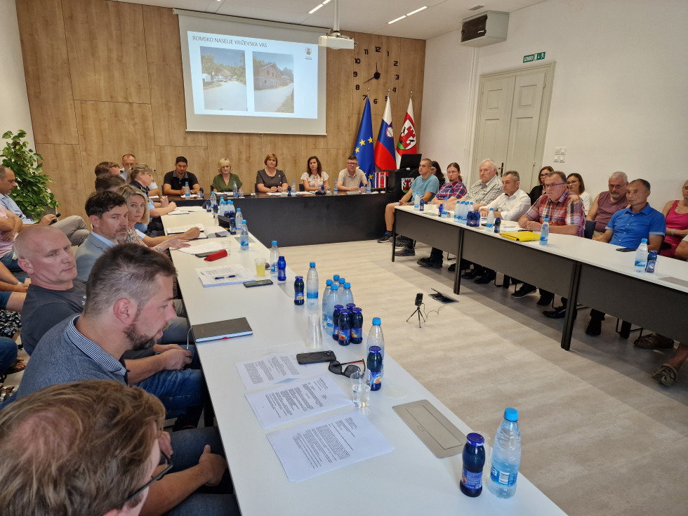 Za mizami sedijo udeleženci sestanka, za njimi lesena stena na kateri je platno s projekcijo, v desnem kotu so zastave
