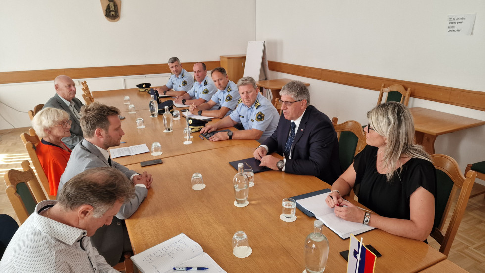 Sodelujoči na sestanku sedijo za dolgo leseno mizo, sedijo nasproti, za njimi je bela stena.