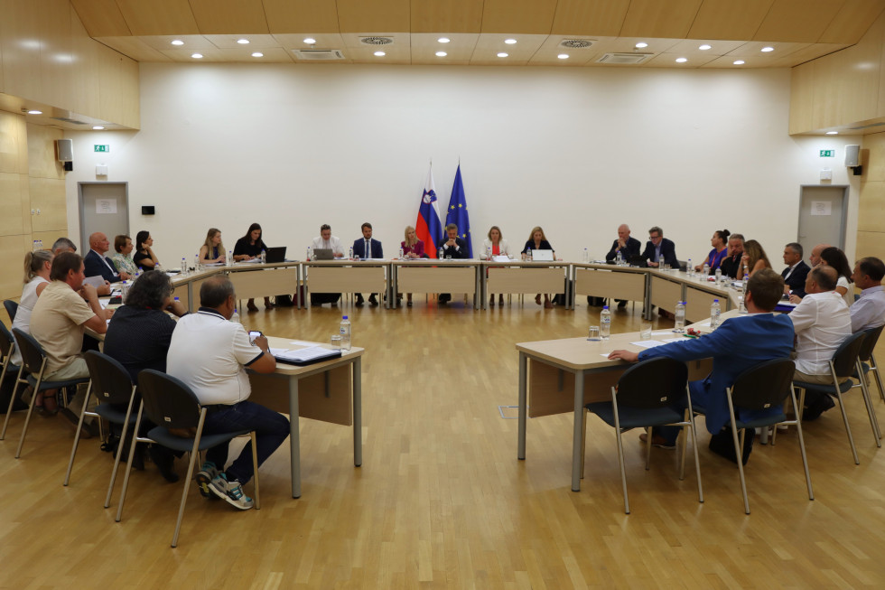 Za mizo, ki je postavljena v krožno formacijo sedijo udeleženci sestanka, za njimi je bela stena in dev zastavi, strop je lesen