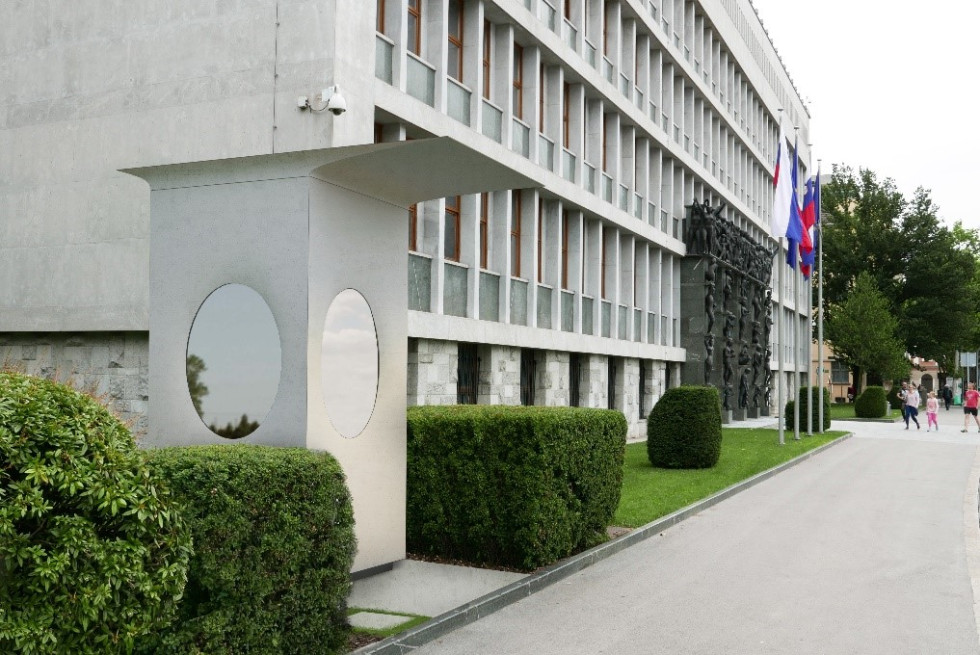 Bela stražarska hiška s steklenimi ovalnimi okni in streho stoji poleg parlamenta, ob njej je živa meja, na levi strani je grm.