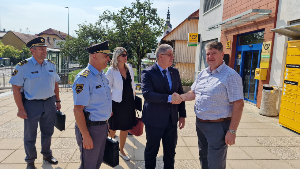 Župan Občine Šentjernej Jože Simončič in minister za notranje zadeve Boštjan Poklukar se rokujeta, na levi strani poleg njiju stojijo ljudje, za njimi na desni strani je stavba pošte