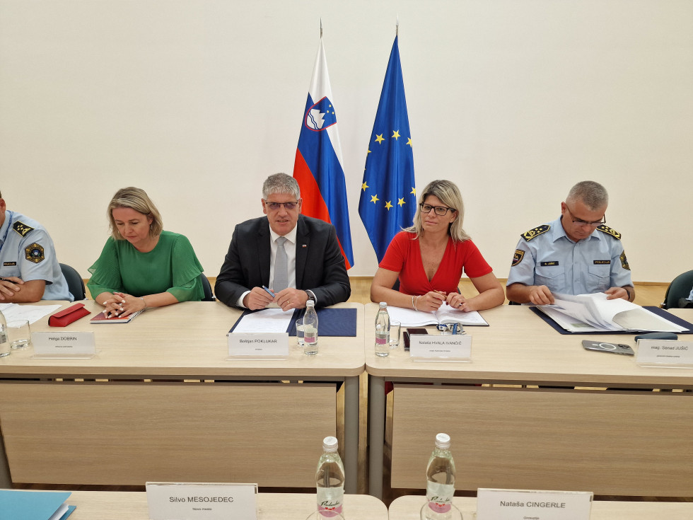 Predstavniki ministrstva za notranje zadeve sedijo za leseno mizo na kateri imajo liste papirja in vodo, za njimi sta postavljeni slovenska in evropska zastava, v ozadju je bela stena.