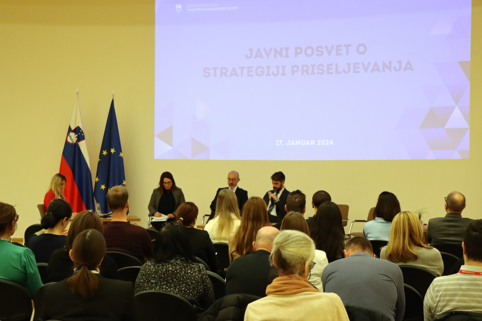 Panelisti in udeleženci javne razprave, na levi strani sta zastavi Slovenije in Evropske unije, na steni je projekcija z naslovom dogodka v modri barvi.