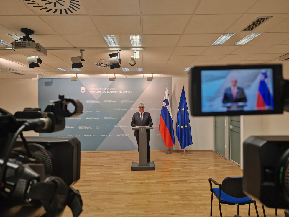 Minister Boštjan Poklukar stoji za sivo govornico. Za njim je modra tabla s celostno grafično podobo ministrstva. za njim sta tudi zastavi Slovenije in Evropske unije. Tla so rjave barve. Pred ministrom so stojijo kamermani, novinarji sedijo na stolih.