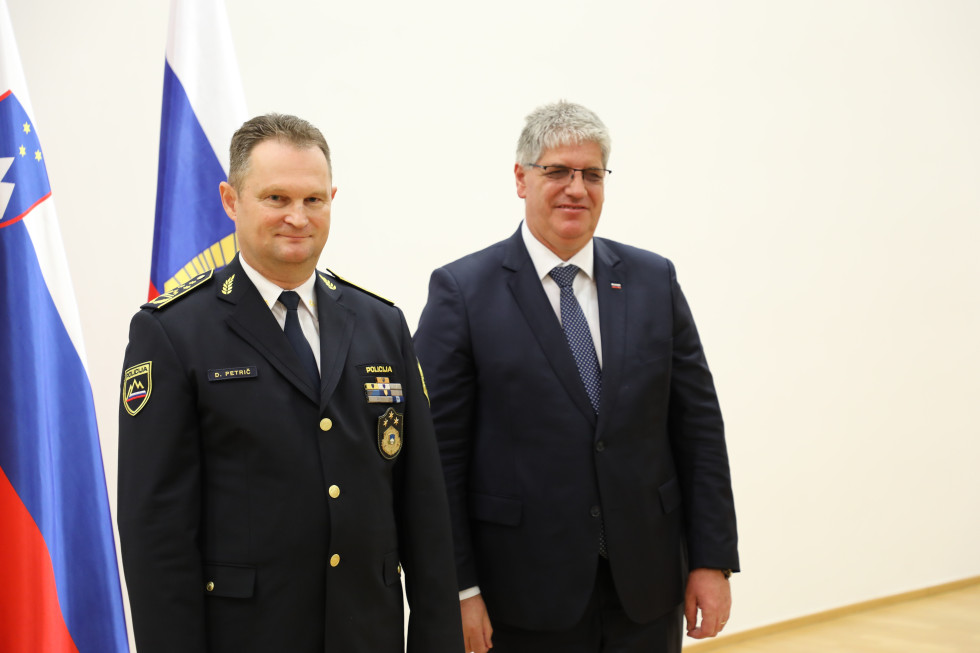 Generalni direktor policije in minister stojita. Za njima je bela stena in zastavi Slovenije in Policije.