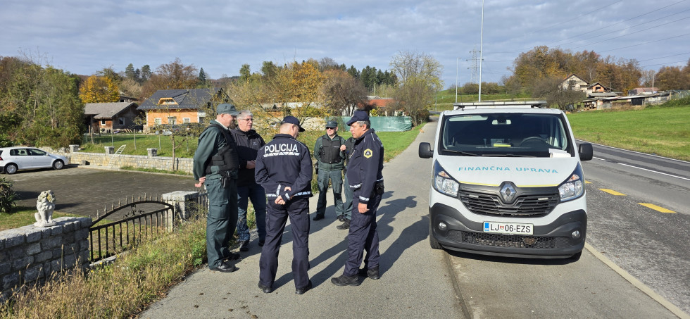 Udeleženci stojijo na pločniku in se pogovarjajo. Poleg njih je parkiran bel kombi z napisom Finančna uprava. Za njimi se vidijo travnik in drevesa.