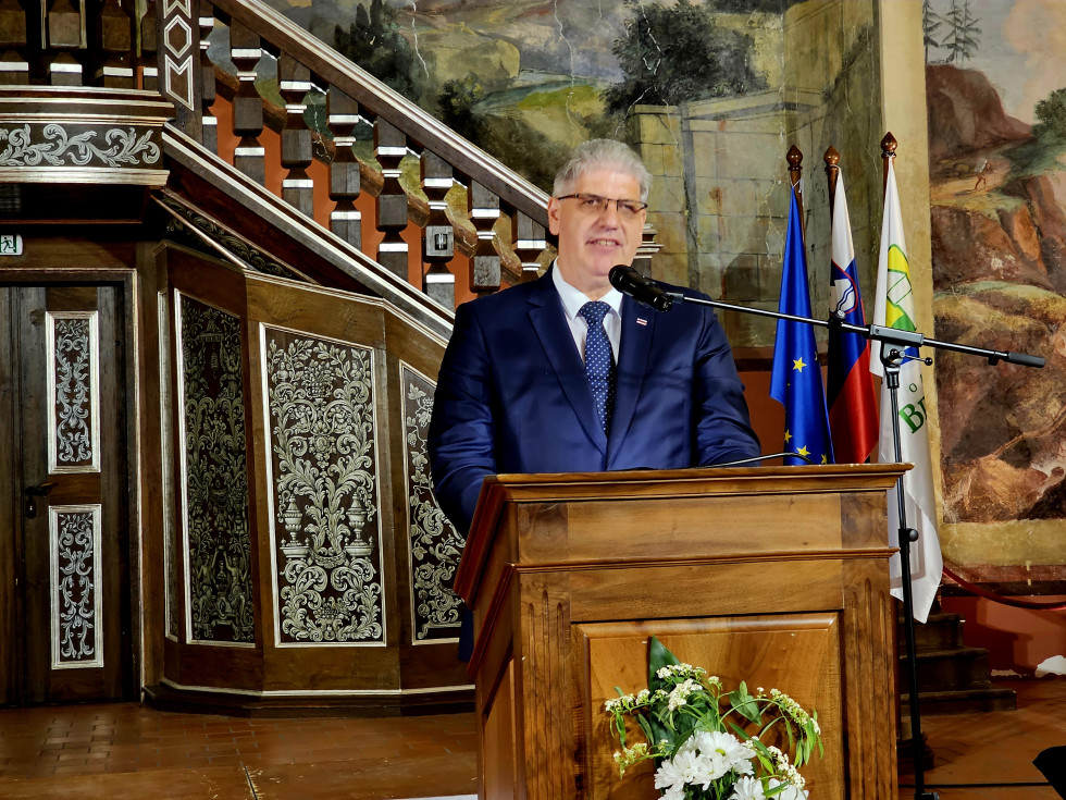 Minister stoji za rjavo govornico. Za njim se vidijo zastave Evropske unije, Slovenije in Občine Brežice.