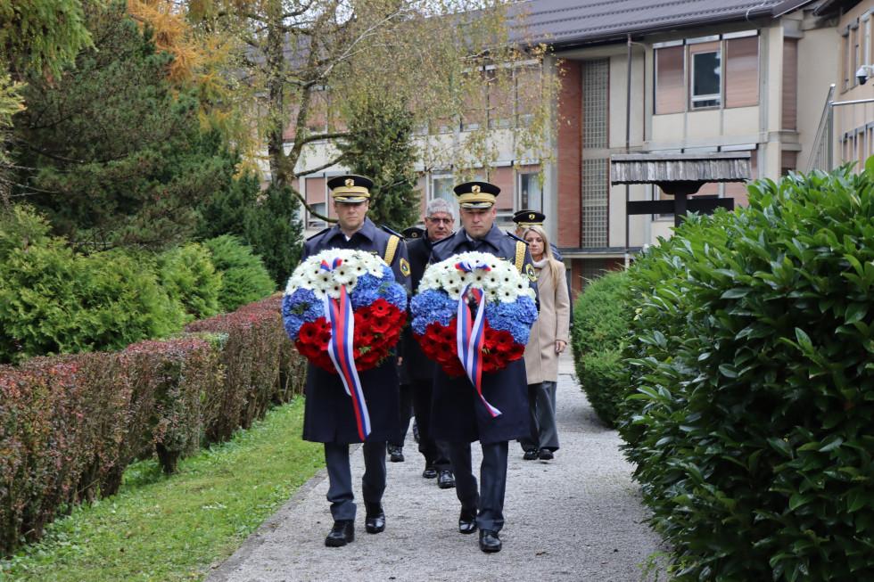 Dva policista nosita dva venca v barvah slovenske zastave. Za njima hodijo člani delegacije. Na tleh so sivi tlakovci, okrog so drevesa brez listov in zelena živa meja. Oblačno.