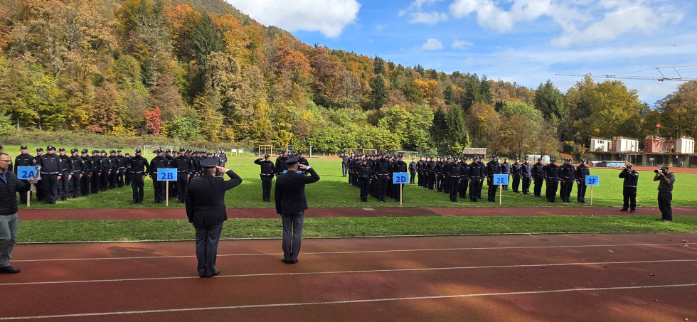 Policistke in policisti stojijo v kolonah na stadionu. Za njimi so zelena drevesa. Pred njimi stojita policista v uniformi, ki jim salutirata.