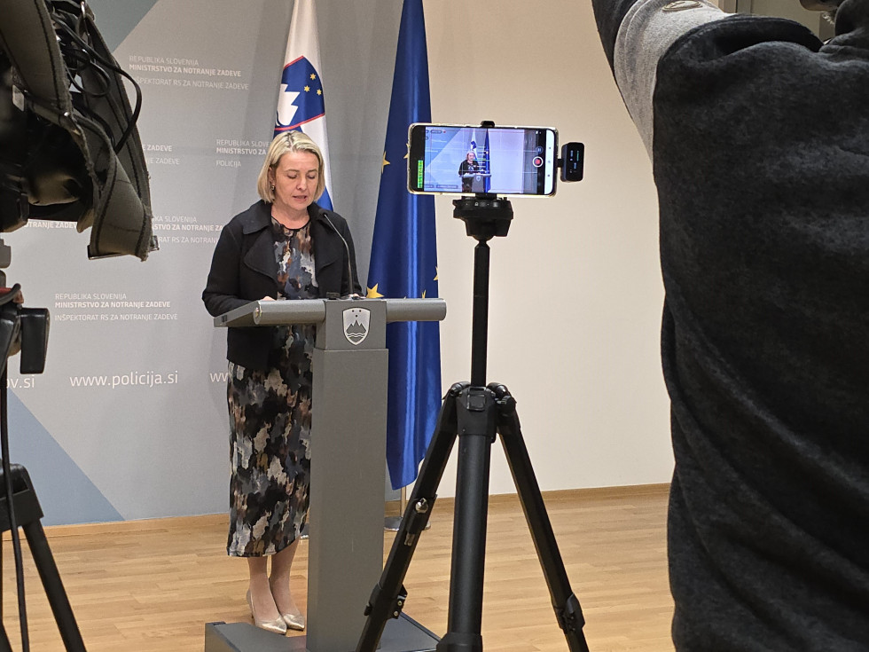 Državna sekretarka stoji za sivo govornico. Za njo stojita zastavi Slovenije in Evropske unije. Del stene za njo je bele barve, del modre. Tla so svetlo rjava. Pred državno stoji črn mikrofon, na katerem stoji mobitel, ki snema izjavo.