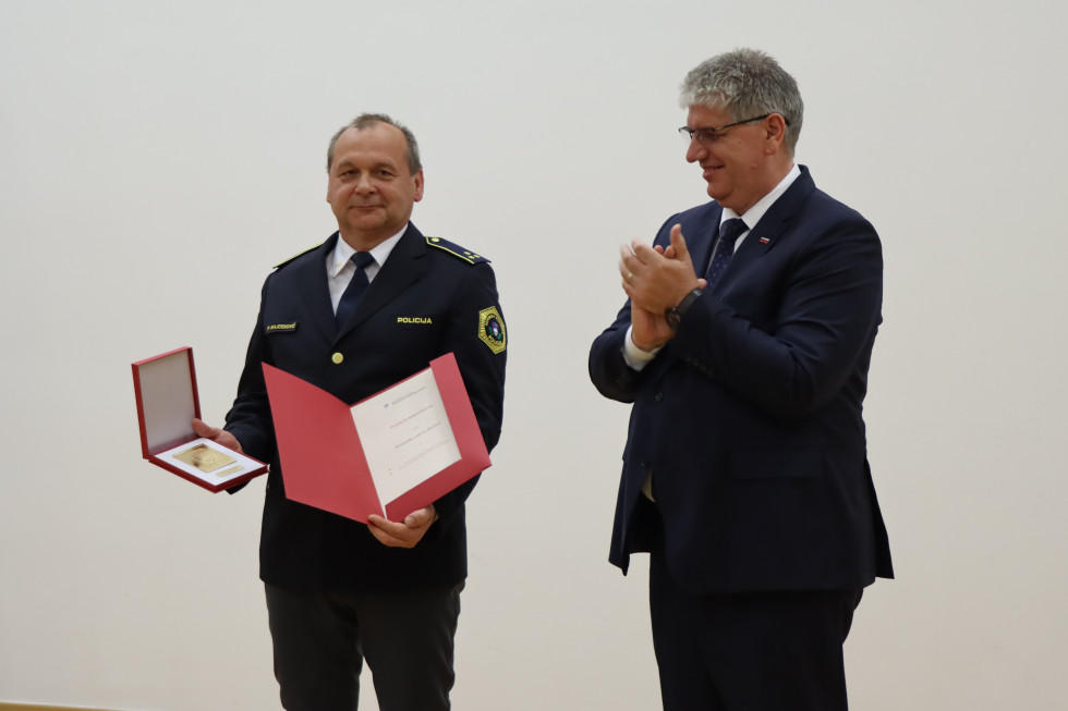 Stojita pred belim ozadjem. Policist ima v roki rdečo mapo in rdečo škatlico z zlato plaketo. Minister ploska.