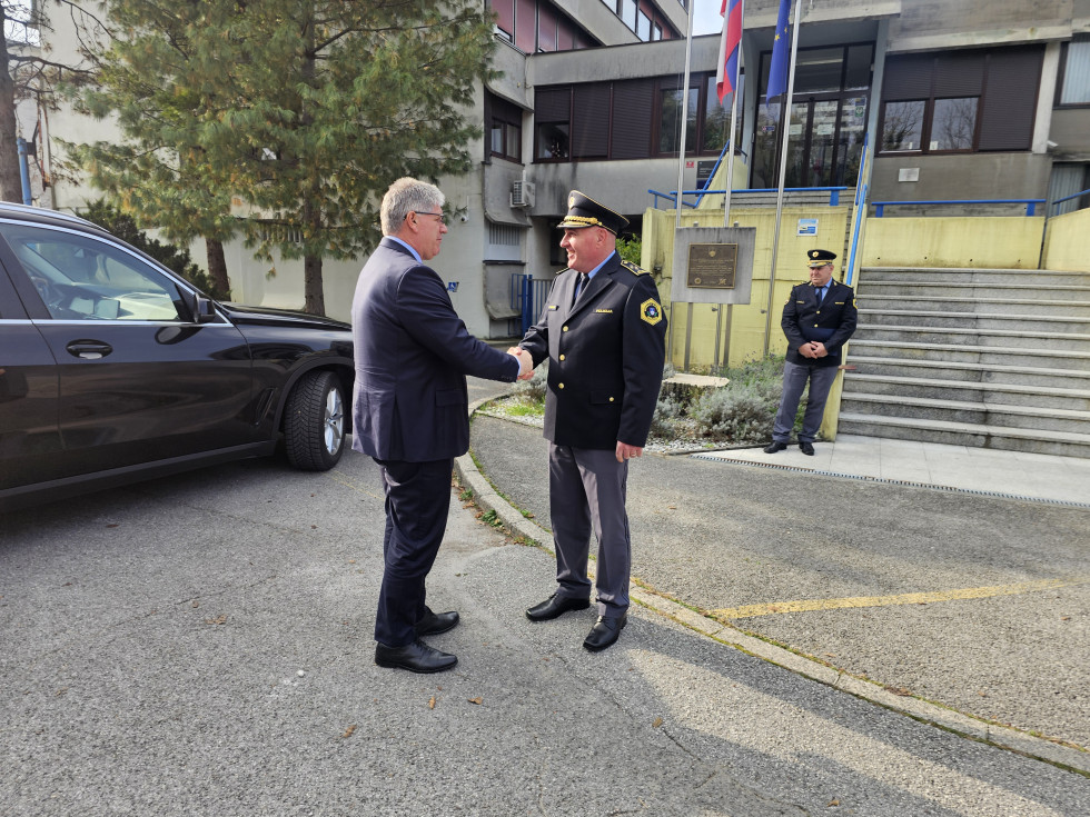 Stojita na asfaltu drug nasproti drugega in se rokujeta. Zadaj je avto in zeleno drevo, desno se vidi del stavbe.