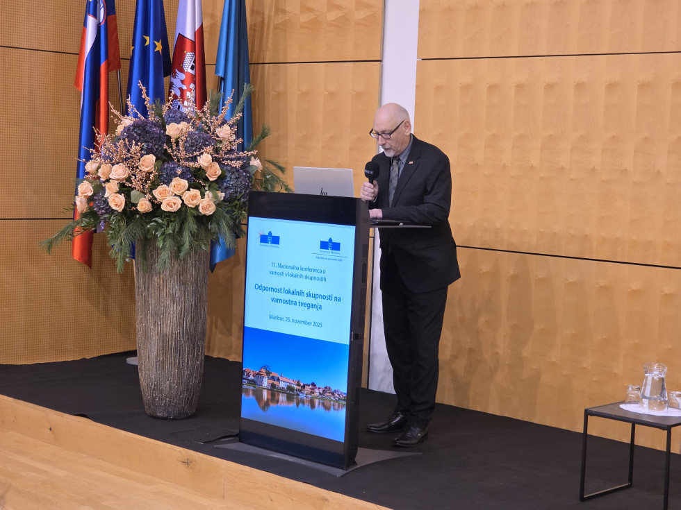 Stoji v dvorani za govornico in govori. Dvorana je v svetlo rjavih tonih. Na levi so zastave in velik cvetlični aranžma. Pred govornico je pokončen ekran.