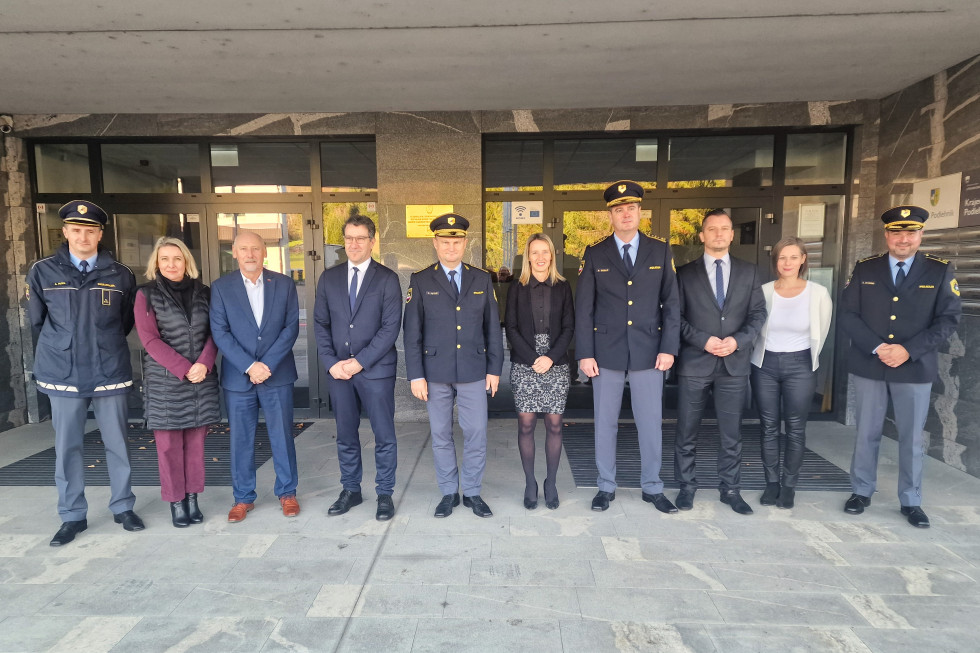 Okrog 10 ljudi stoji pred vhodom v stavbo. Tla so tlakovana in sive barve.