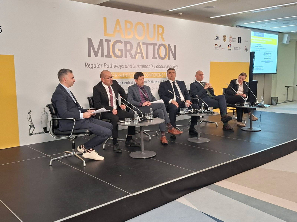 Udeleženci sedijo na stolih, ki stojijo na črnem odru. Za njimi je bel pano, na katerem v angleščini piše delovne migracije.