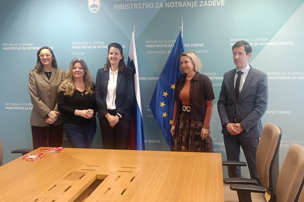 Stojijo v sejni sobi, za svetlo rjavo mizo, za njimi je svetlo moder zaslon z belimi napisi ter slovenske ter evropska zastava.