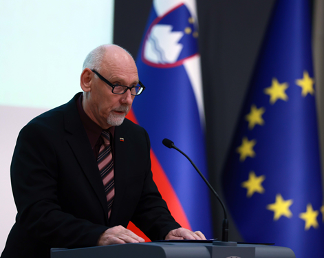 Minister stoji za sivo govornico, na kateri drži obe roki. Za njim se vidita zastavi Slovenije in Evropske unije.