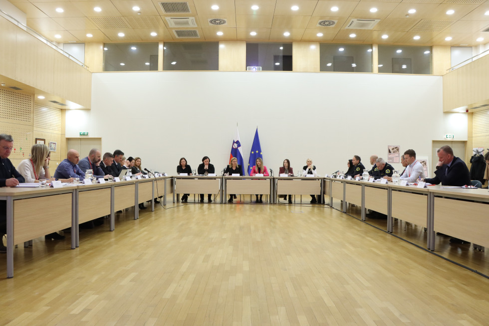 Udeleženci sedijo za mizami, postavljenimi v obliki črke U, za njimi je bela stena, ob njej pa slovenska in evropska zastava