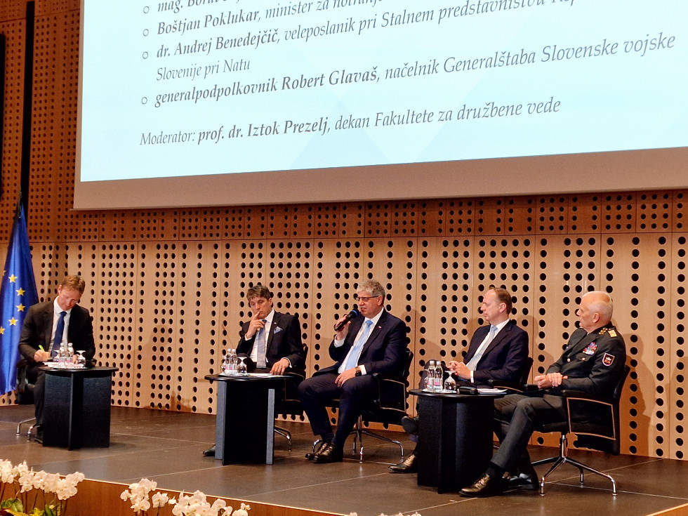 Štirje panelisti sedijo na črnih stolih. Stena za njimi je v rjavih tonih.