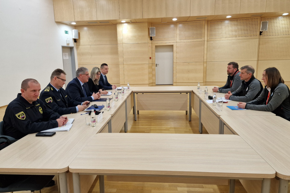 Osebe si sedijo nasproti, za mizami, ki so postavljene v pravokotnik. Mizo so svetlo rjave, prav tako dvorana. Na mizah so kozarci in steklenice z vodo.