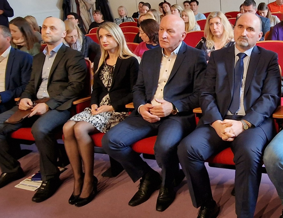 Udeleženci sedijo drug ob drugem na rdeči stolih v več vrstah in poslušajo