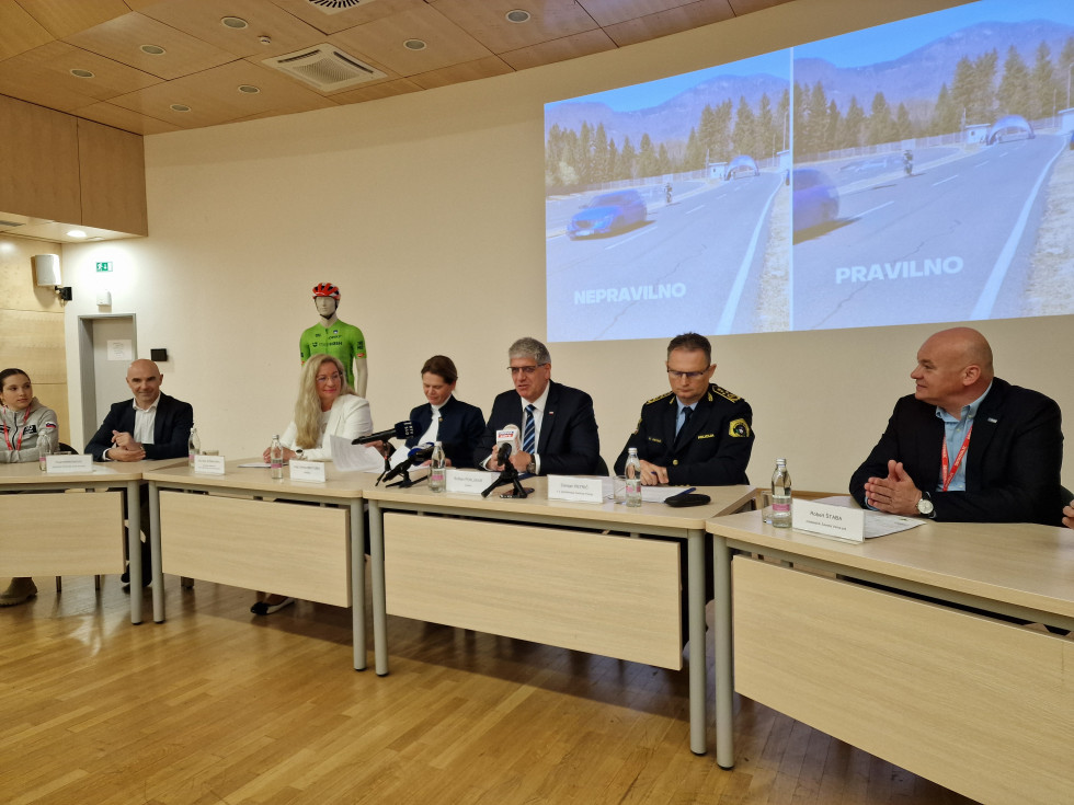 Govorci sedijo za svetlo rjavimi mizami v svetlo rjavi dvorani. Na tleh je parket. Za govorci je projekcija s prikazom prehitevanja kolesarja. Levo je lutka kolesarja v zelenem dresu in z rdečo čelado na glavi.