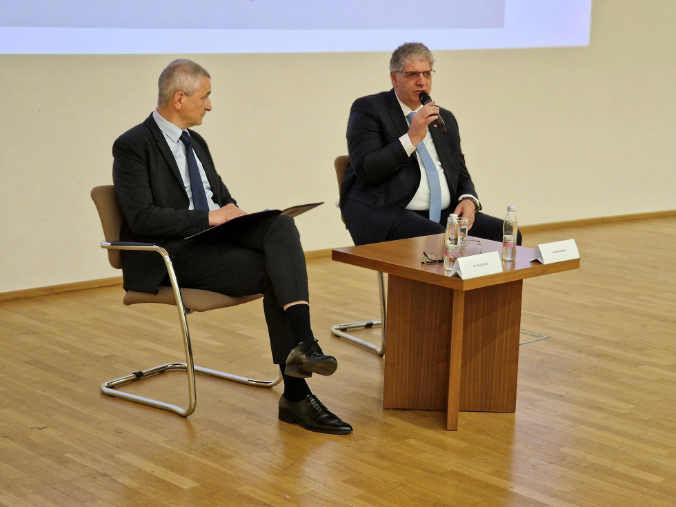 Govorca sedita na stolih, eden od njiju drži v roki mikrofon. Pred njima je nizka kvadratna mizica rjave barve, tudi tla so svetlo rjave barve.