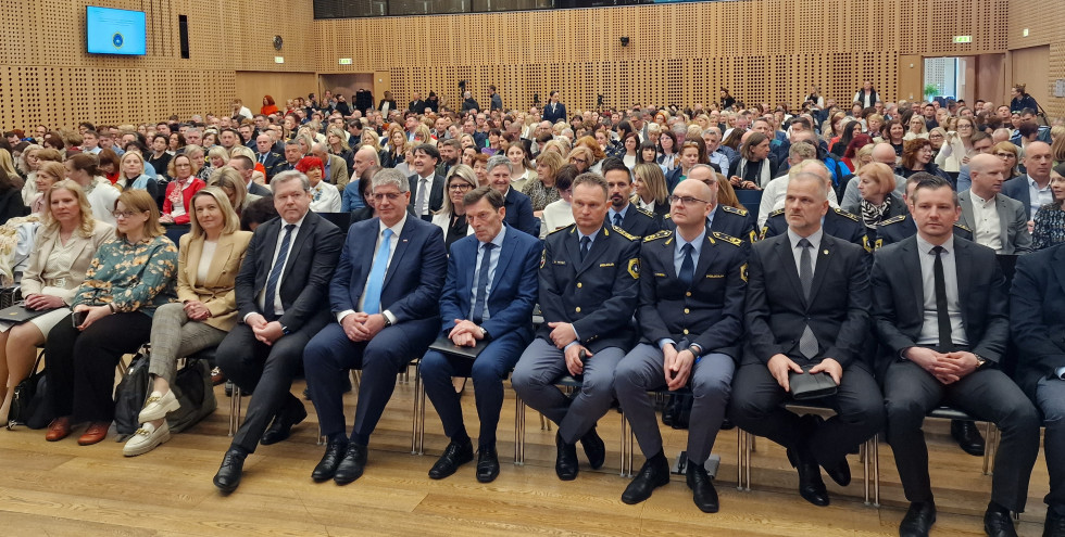 Ljudje sedijo v veliko vrstah v polni dvorani in gledajo proti odru