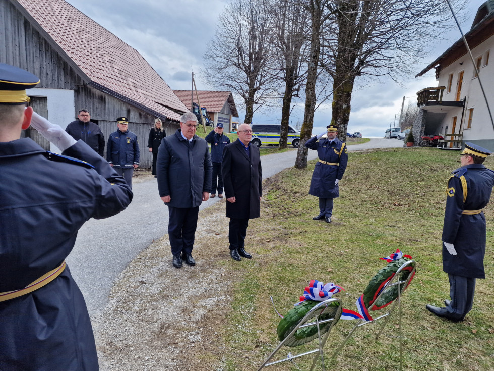 Dva moška stojita na prostem in spoštljivo gledata predse. V ospredju sta vidna dva spominska venca na stojalih. V ozadju je nekaj ljudi.