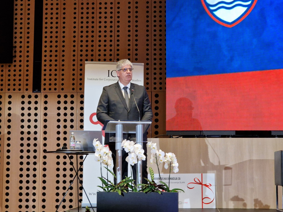 Stoji na odru za govornico. Desno od njega je projekcija na platnu z veliko slovensko zastavo.