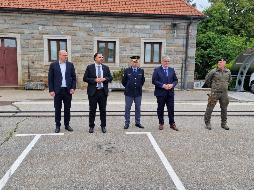 Možje stojijo na prostem, na asfaltu. Za njimi je hiša s kamnito fasado in tremi okni.