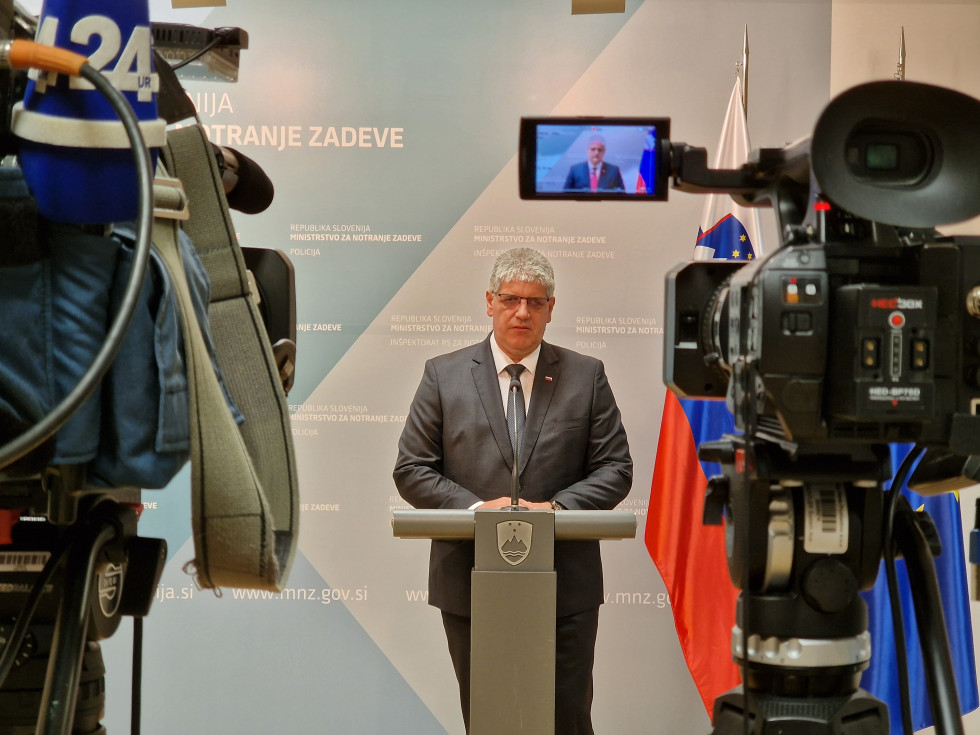Minister stoji za sivo govornico. Za njim je ozadje sivo-modre barve ter stojalo s slovensko zastavo. Na levi in desni strani fotografije se vidita kameri, ki snemata ministrovo izjavo.