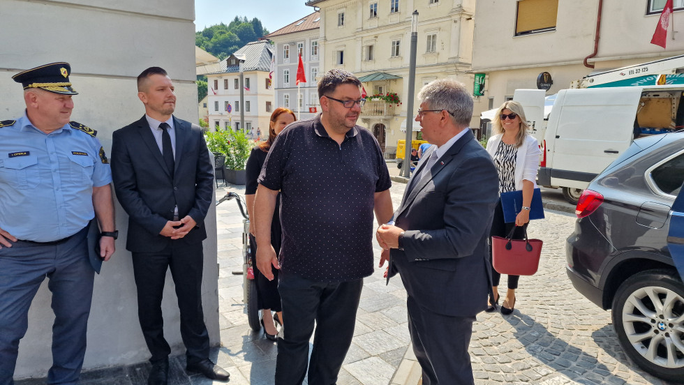 Stojita na prostem v mestu. Dan je sončen, okoli njiju so še druge osebe.