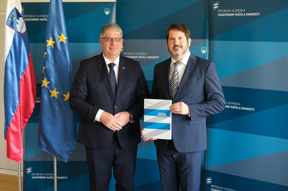 Minister Boštjan Poklukar in zagovornik načela enakosti Miha Lobnik stojita pred modro steno, kjer se vidi zastava Evropske unije. Gospod Lobnik v roki drži knjigo - letno poročilo o delu zagovornika v 2024.